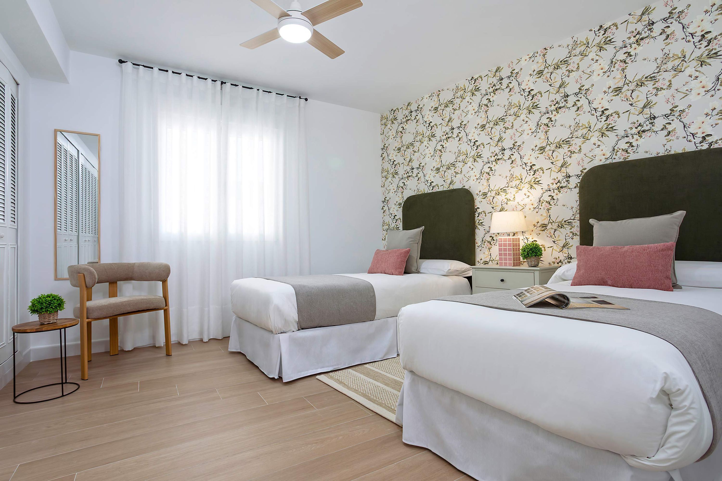 Une chambre d’hôtel avec deux lits, une chaise, une petite table avec une plante et un ventilateur de plafond.