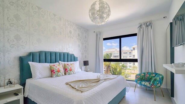 A bedroom with a large bed, a nightstand, a chair, a window with curtains, and a view of buildings outside.