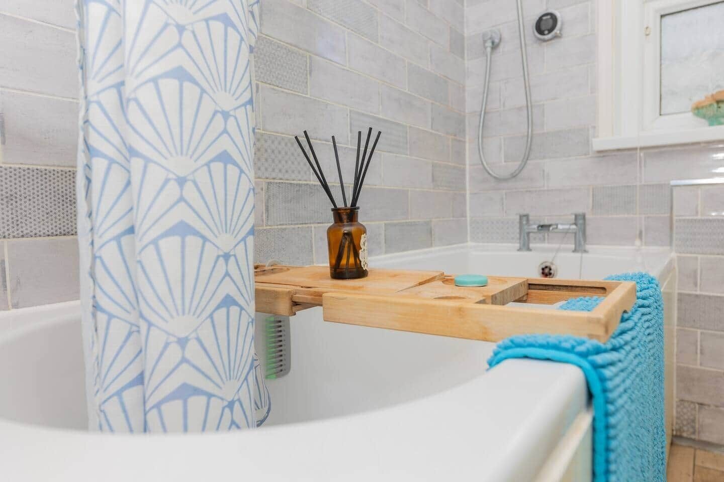 A bathroom with a bathtub, a wooden shelf with a bottle and straws, a blue towel, and a shower area.