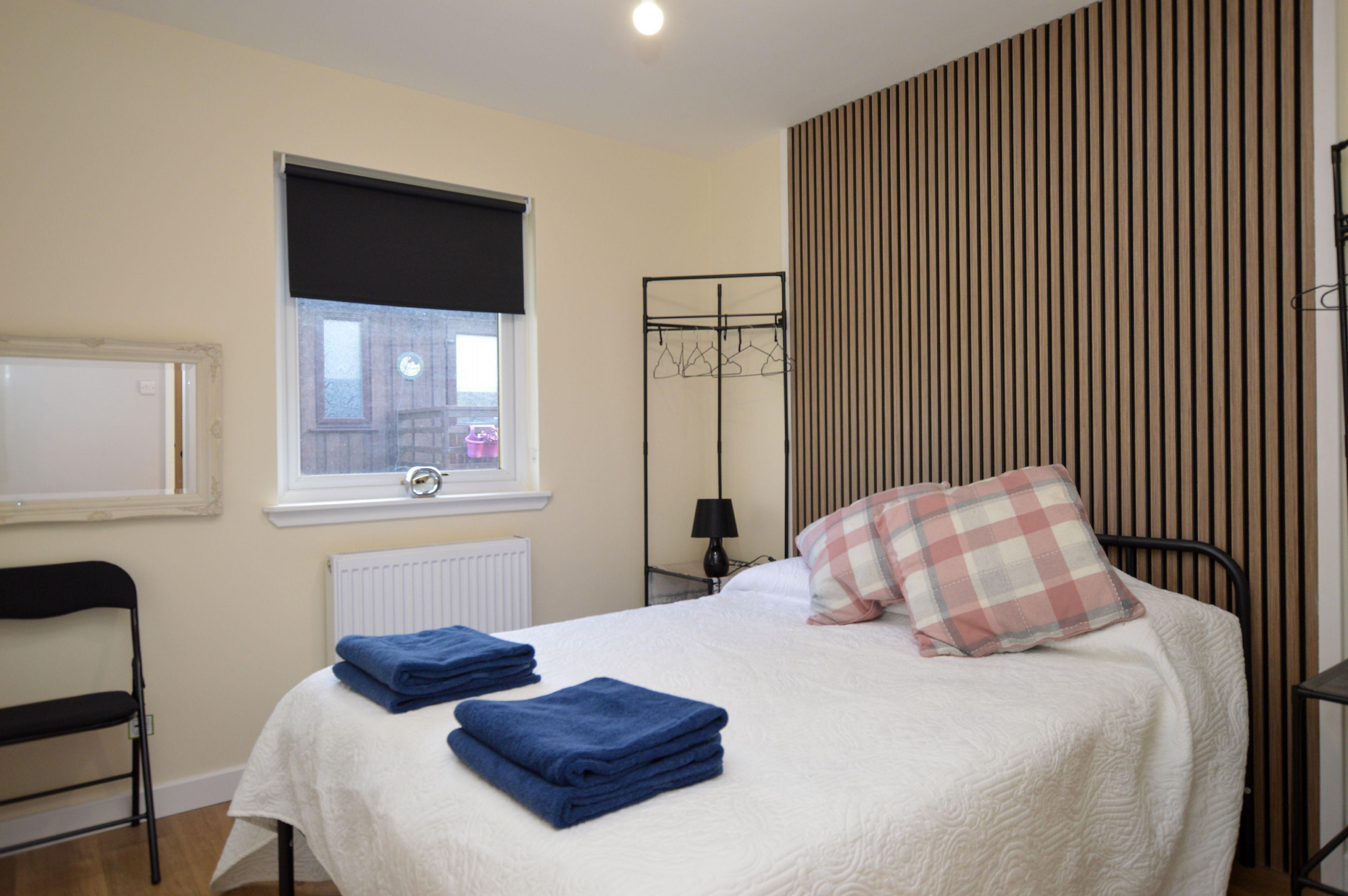 A bedroom with a bed, a radiator, a window with a blind, a coat rack, and a chair.
