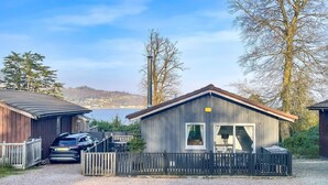 Eine Holzhütte mit Kiesweg, einem davor geparkten Auto und Seeblick mit Hügeln im Hintergrund.