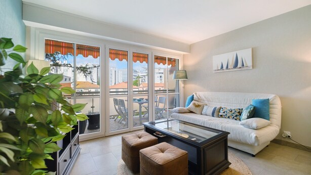 A living room with a sofa, ottomans, a coffee table, a lamp, and a view through large glass doors. - Juan-les-Pins Vitoria Grand 2P – Quiet, Parking, Central Location (Antibes)