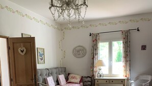 A bedroom with a bed, a chair, a dresser, a window with curtains, a chandelier, and a door. - Charming bed and breakfast in Giono country. (Manosque)