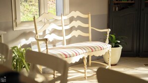 A white wooden bench with a striped cushion, a potted plant, and a window with a view of greenery. - Charming bed and breakfast in Giono country. (Manosque)