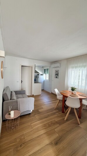 A modern living room with a grey sofa, a round wooden dining table with white chairs, a small side table, and a kitchen in the background. - House Mar Salou (Salou)