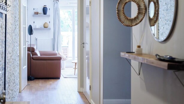 Un couloir avec un plancher en bois, un banc, un canapé et une table à manger. Il y a deux miroirs au mur et un luminaire au plafond.