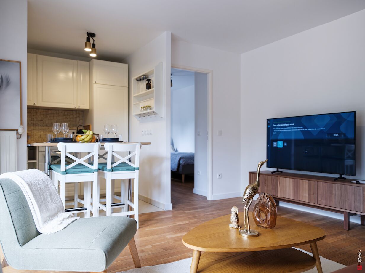 Un salon moderne avec un canapé, une table basse et un téléviseur. On y trouve également un coin repas avec une table et des chaises, et une cuisine à l’arrière-plan.