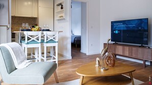 Un salon moderne avec un canapé, une table basse et un téléviseur. On y trouve également un coin repas avec une table et des chaises, et une cuisine à l’arrière-plan.