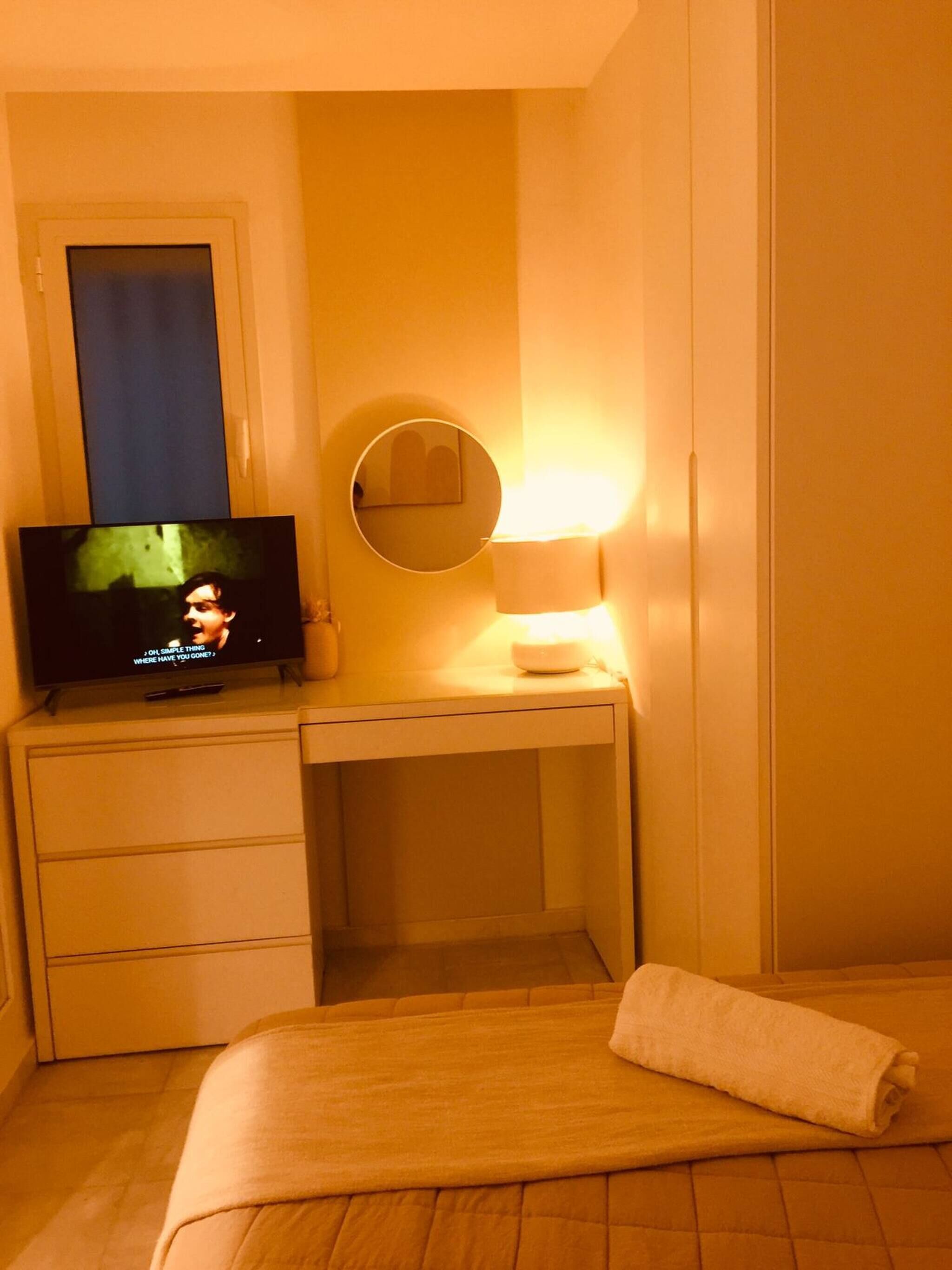 A hotel room with a bed, a desk with a television, a mirror, and a bedside lamp.