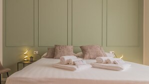 Un lit bien fait, avec des serviettes pliées en rouleaux, et une petite table sur laquelle se trouvent une lampe et une plante.