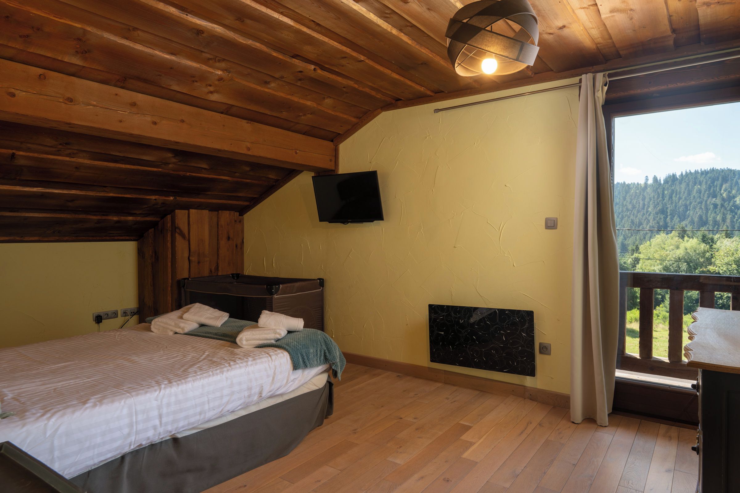 Una camera da letto con soffitto in legno, un letto con coperta verde, una TV a schermo piatto montata a parete e una finestra vista foresta.