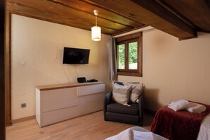 A bedroom with a wooden ceiling, a TV mounted on the wall, a bedside table, a chair, and a window. - Chalet Serenity - Sleeps 9 - Hot Tub (Liézey)