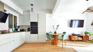 A modern kitchen with white cabinets, a dining area with green chairs, a round wooden table, and a flat-screen TV mounted on the wall. - 2 bedrooms. Roman Amphitheater. Near the beach (Tarragona)