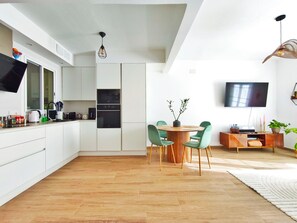 Eine moderne Küche mit weißen Schränken, einem Essbereich mit grünen Stühlen, einem runden Holztisch und einem an der Wand befestigten Flachbildfernseher.