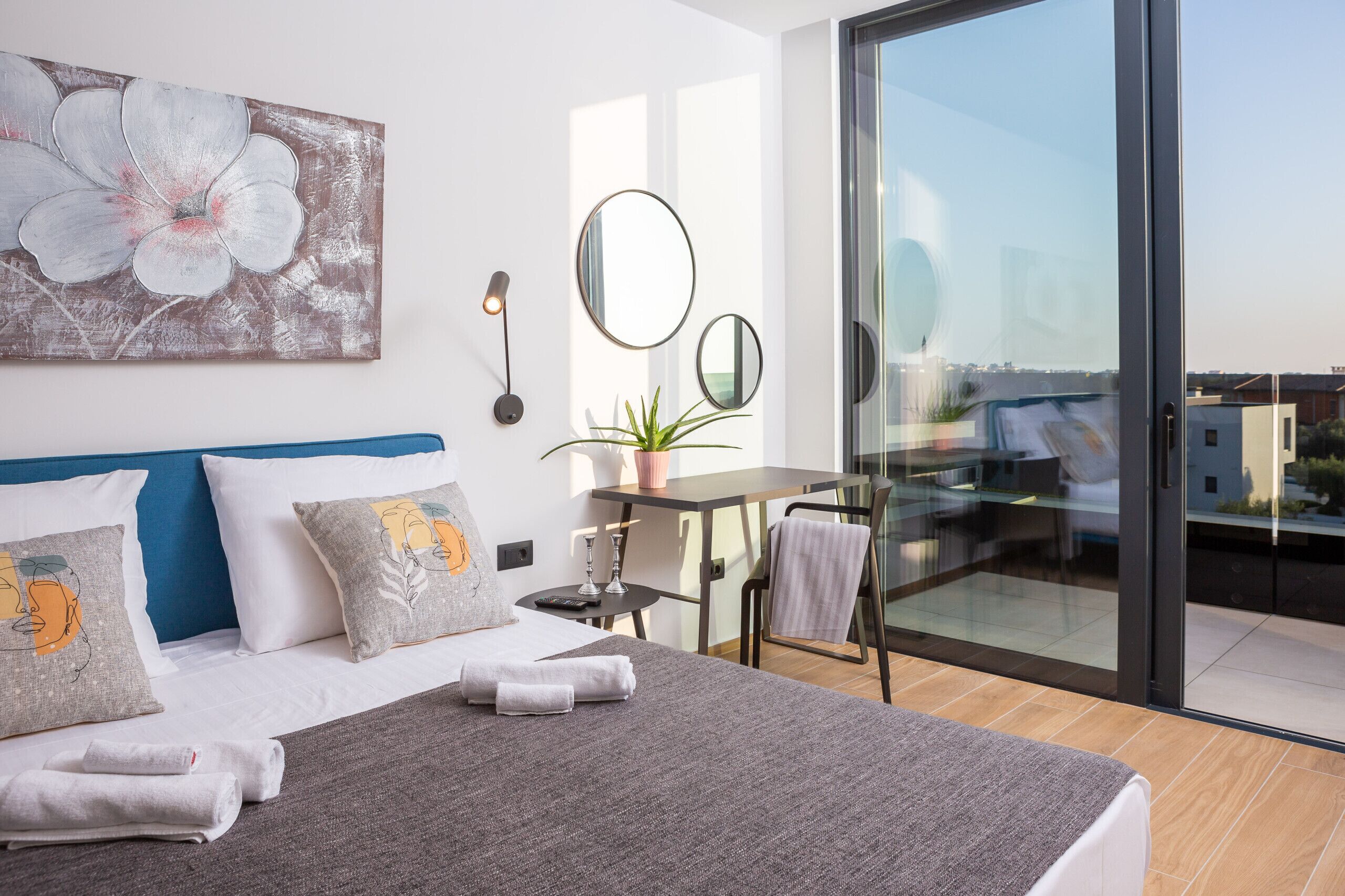 Moderni hotellihuone, jossa on suuri sänky, työpöytä, tuoli, ruukkukasvi ja parveke, jonka ovena on lasiovi.