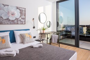 Ein modernes Hotelzimmer mit einem großen Bett, einem Schreibtisch, einem Sessel, einer Topfpflanze und einem Balkon mit Glastür.