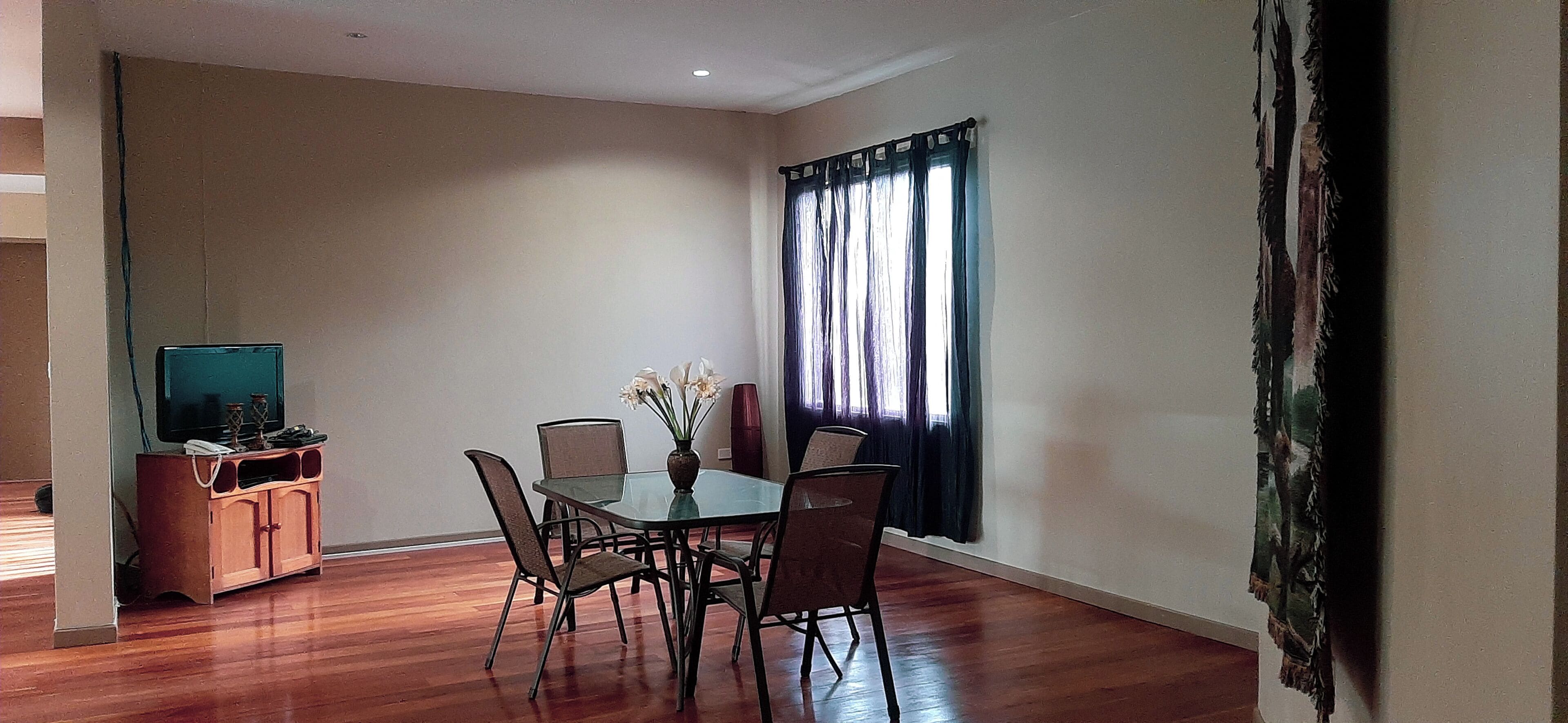 A dining area with a glass table, six chairs, a vase with flowers, a television on a wooden cabinet, and a window with curtains.