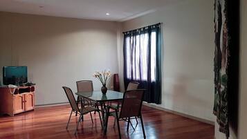 A dining area with a glass table, six chairs, a vase with flowers, a television on a wooden cabinet, and a window with curtains.