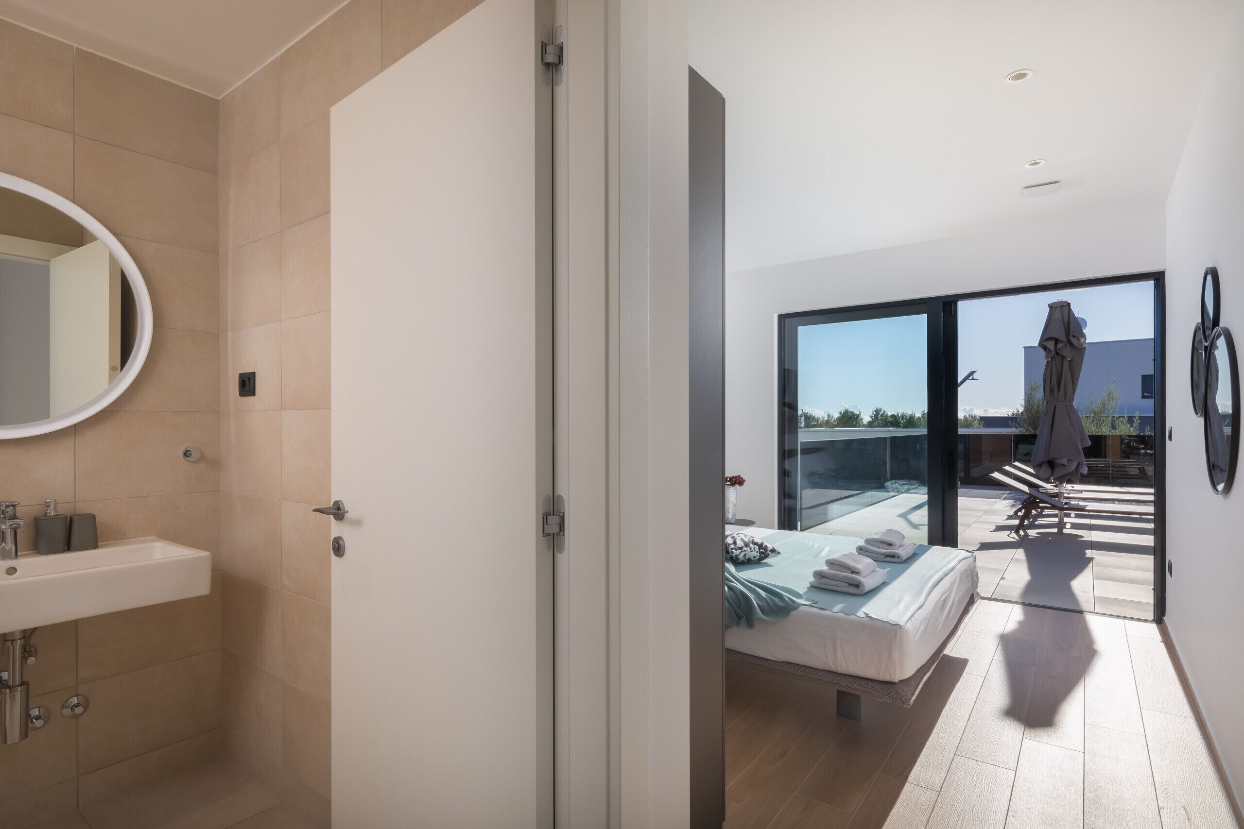 Une chambre à coucher moderne avec un grand lit, une salle de bain avec un lavabo et une vue sur l’extérieur grâce à une porte vitrée.