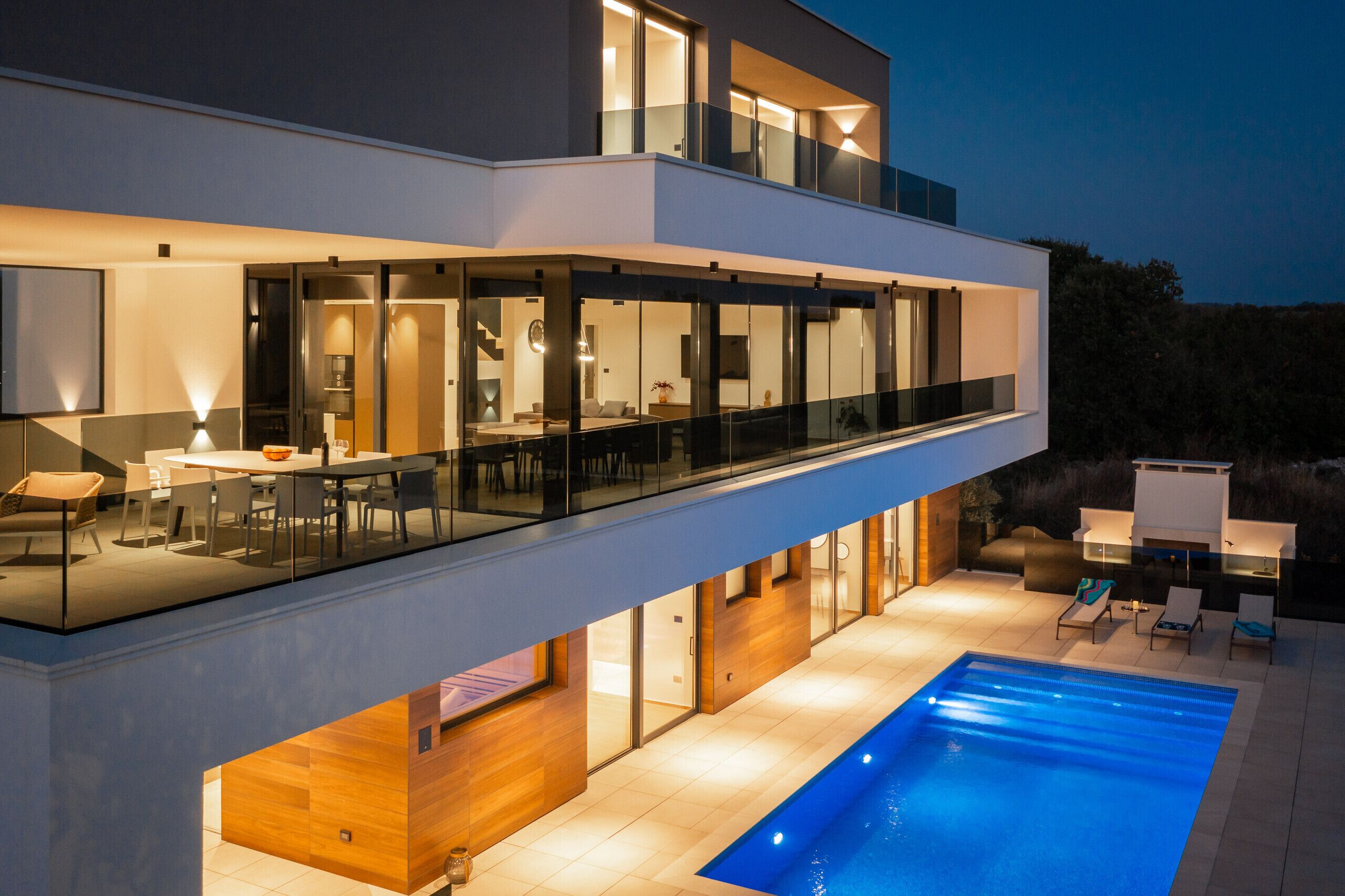 A modern two-story house with a glass balcony, a swimming pool, and a well-lit interior.