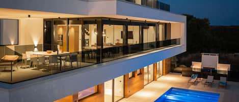 Uma casa moderna de dois andares com varanda de vidro, piscina e interior bem iluminado.