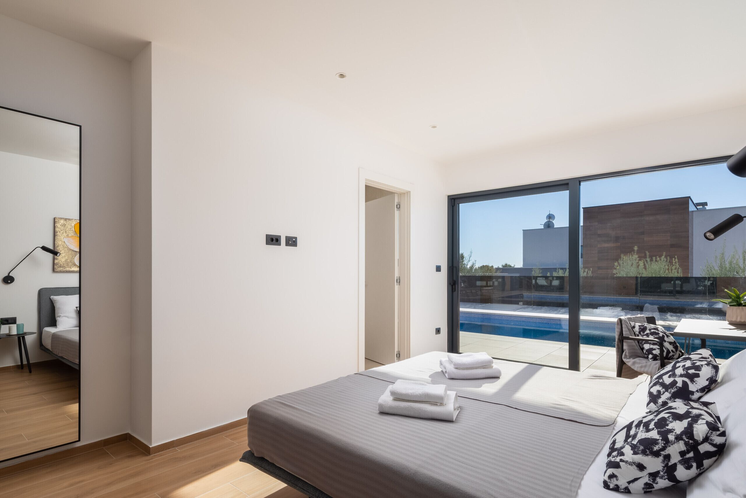 A modern bedroom with a large bed, a view of a swimming pool, and a door leading to another room.