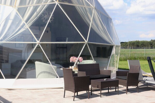 Une structure en forme de dôme géodésique, dotée d'une façade en verre, de meubles en osier et offrant une vue sur un paysage verdoyant.