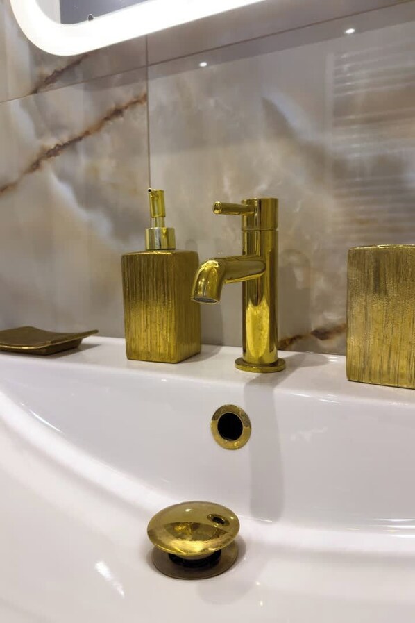 A bathroom with a white sink, gold faucets, and a marble backsplash.