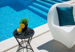 Ein Poolbereich mit einer weißen Liege, einem kleinen runden Tisch mit einer Pflanze, einem Glas Orangensaft und einer Sonnenbrille.