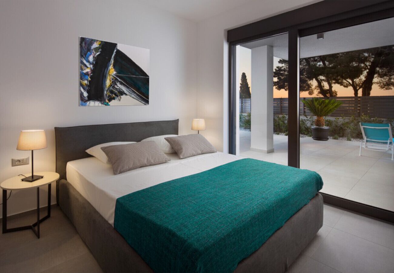 A modern hotel room with a large bed, a painting on the wall, a small table with a lamp, and a view of the outdoors.
