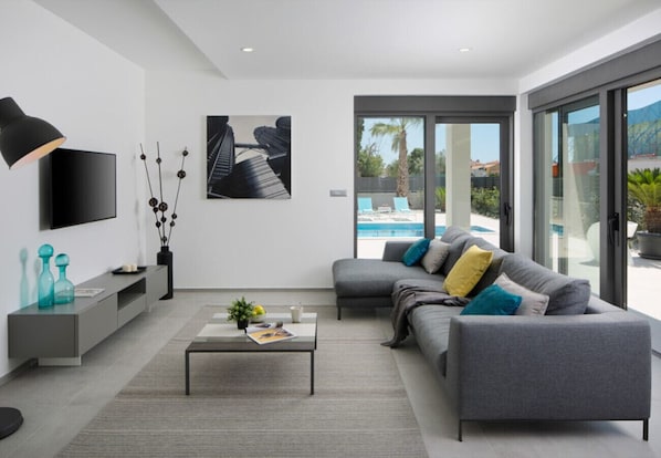 Ein modernes Wohnzimmer mit grauem Sofa, einem Couchtisch, einem Fernseher und einem großen Fenster mit Blick auf einen Pool.