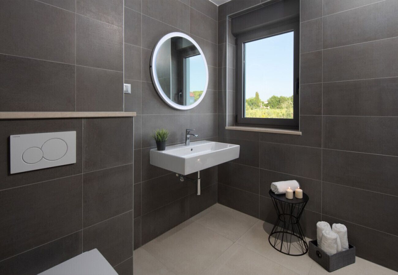 Une salle de bain moderne avec un lavabo blanc, un miroir rond, une fenêtre et une petite table sur laquelle sont posées des serviettes.