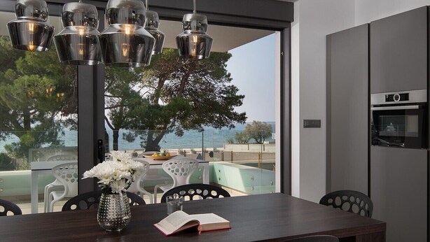 Un espace repas moderne avec une table en bois foncé, des chaises en métal, un vase rempli de fleurs et une vue sur la mer grâce à de grandes fenêtres.