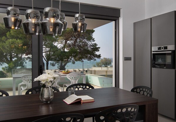 A modern dining area with a dark wooden table, metal chairs, a vase with flowers, and a view of the sea through large windows. - Vivi Rent Fažana - Villa Fasana Penthouse 5 (Fažana)