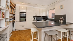 A modern kitchen with white cabinetry, a central island with bar stools, a built-in oven, and a window with a view. - Monkgate Cloisters - Free Parking (York)