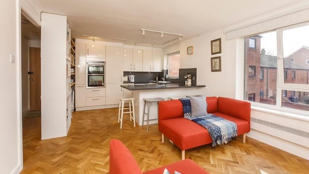 A modern kitchen with white cabinets, a central island, and a red sofa in a bright room with wooden flooring and large windows. - Monkgate Cloisters - Free Parking (York)