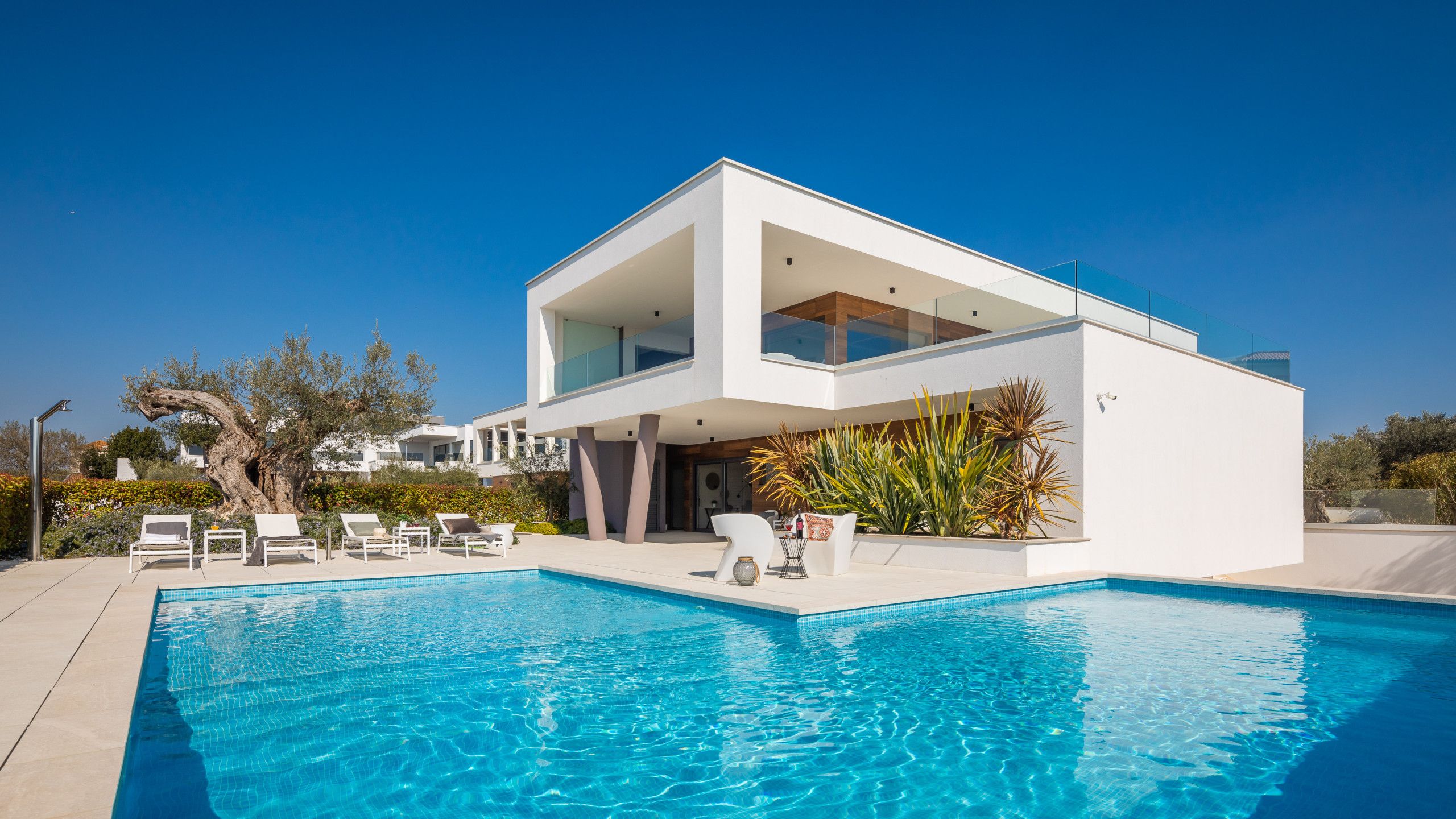 Een modern, tweelaags huis met een zwembad, buitenzitplaatsen en een helderblauwe lucht.