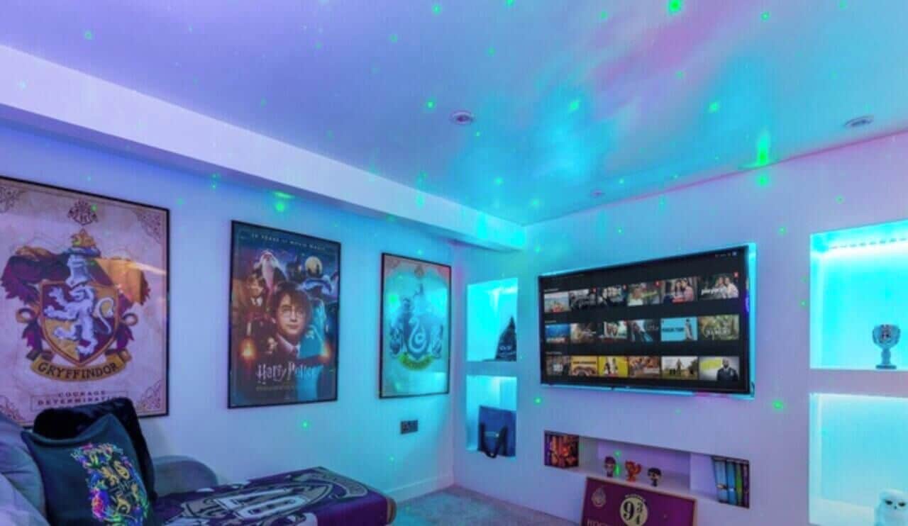Um quarto com uma prateleira na parede exibindo diversos cartazes e recordações, uma cama com um travesseiro temático de Harry Potter e uma luminária no teto.