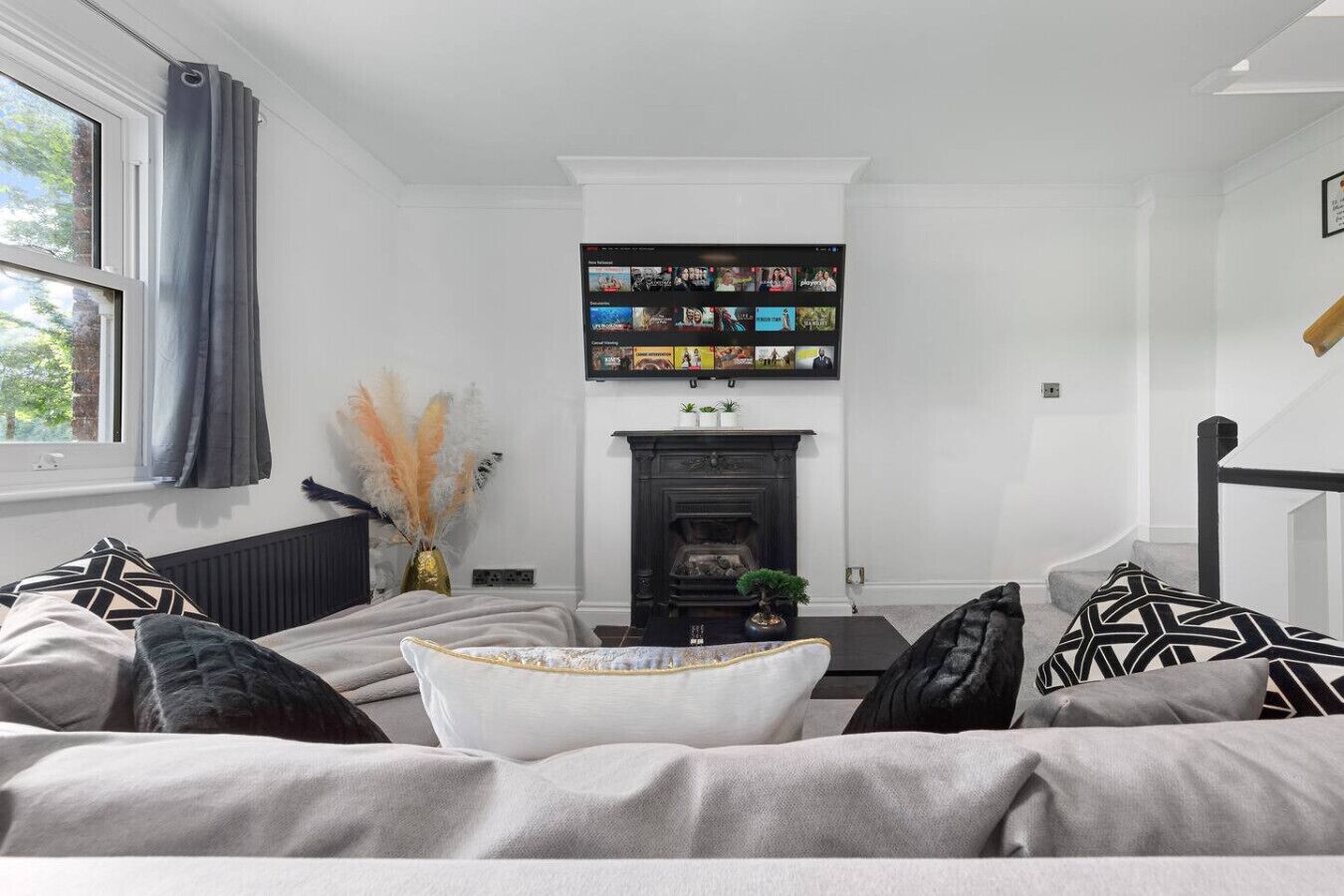 A modern living room with a fireplace, a flat-screen TV mounted above it, and a comfortable seating area with patterned cushions.