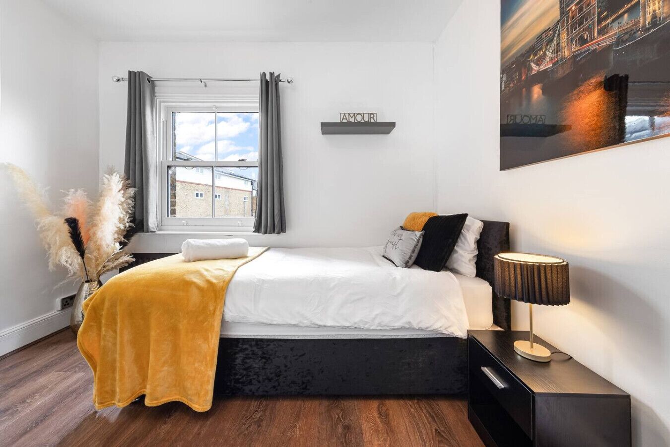 A modern bedroom with a bed, a nightstand, a lamp, a window with curtains, a wall-mounted shelf with a sign, and a cityscape painting.