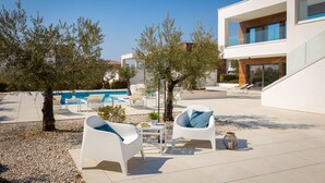 Patio esterno moderno con arredi bianchi, piscina e un edificio con ampie finestre.
