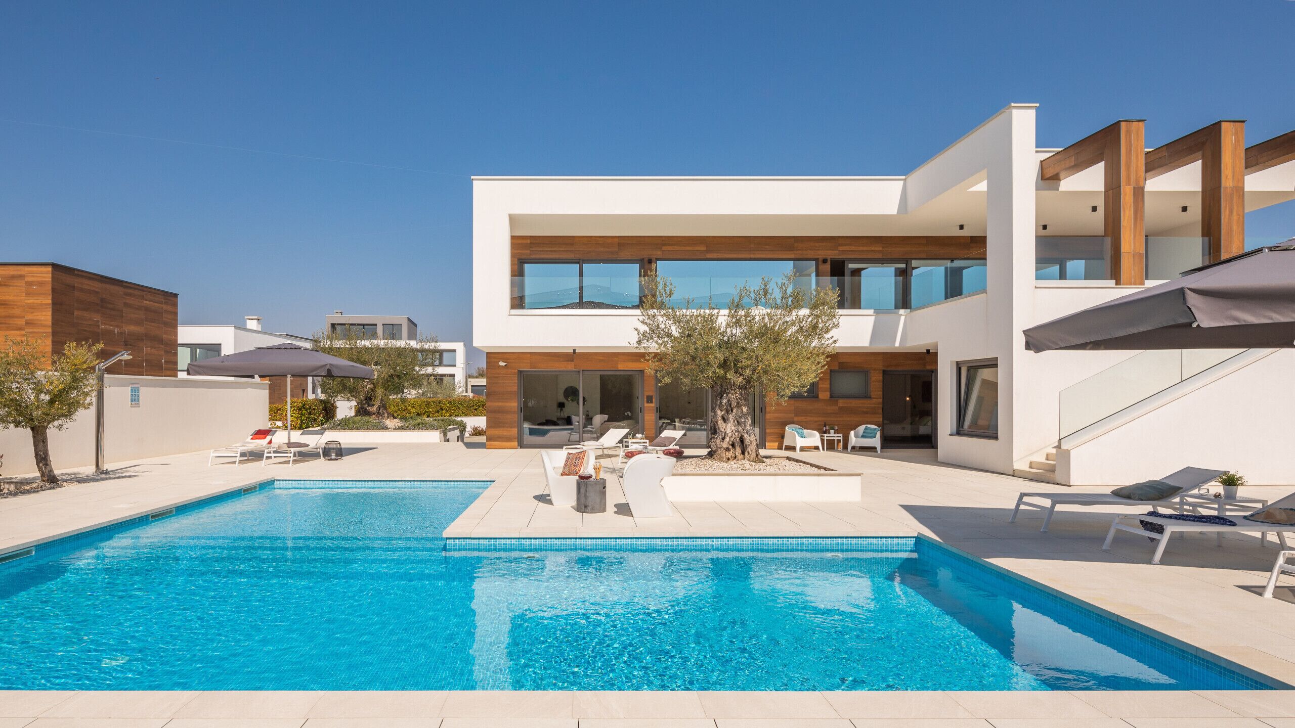 Un espace moderne aménagé au bord de la piscine, avec une piscine à l’eau d’un bleu limpide, des chaises longues et un bâtiment contemporain doté de grandes fenêtres.