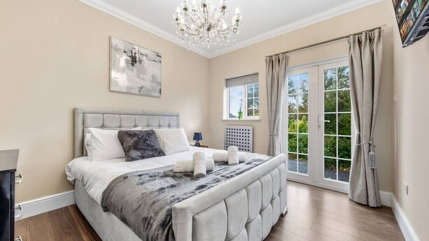 Ein Schlafzimmer mit einem großen Bett, einer Kristallleuchtre, einem Fenster mit Vorhängen und Blick ins Grüne.