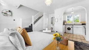 Ein Schlafzimmer mit einem Bett, einem Holztisch, einer Vase mit Blumen, einer Treppe und einer Küchenzeile mit einer Waschmaschine.