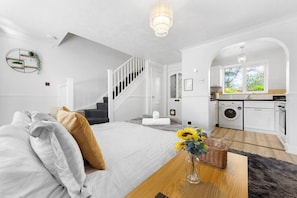 Ein Schlafzimmer mit einem Bett, einem Holztisch, einer Vase mit Blumen, einer Treppe und einer Küchenzeile mit einer Waschmaschine.