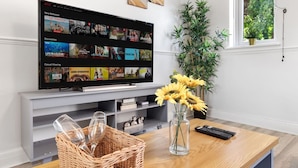 Ein Wohnzimmer mit einem Flachbildfernseher, der eine Streaming-Dienstanzeige zeigt, einem Holztisch mit einer Vase voller Sonnenblumen, einem Korbwäschesessel und einer Fernbedienung.