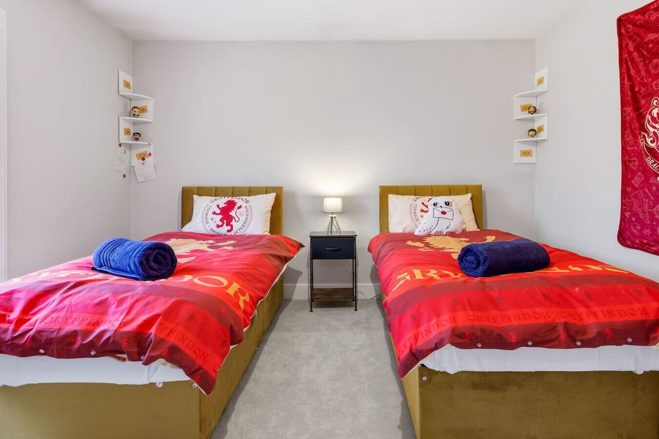 Een tweepersoonskamer met twee bedden, elk voorzien van een rode deken, een nachtkastje en een lamp, plus een rode wandtapijt aan de muur.