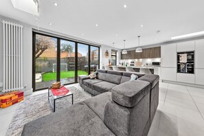 A modern living room with a grey sectional sofa, a glass door leading to a garden, and a kitchen with white cabinets and a built-in oven. - The Lumos House, 4 Bed property W Free Parking (London)