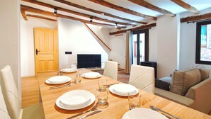 Uma sala de jantar com uma mesa de madeira posta para quatro pessoas, uma televisão e um sofá ao fundo.