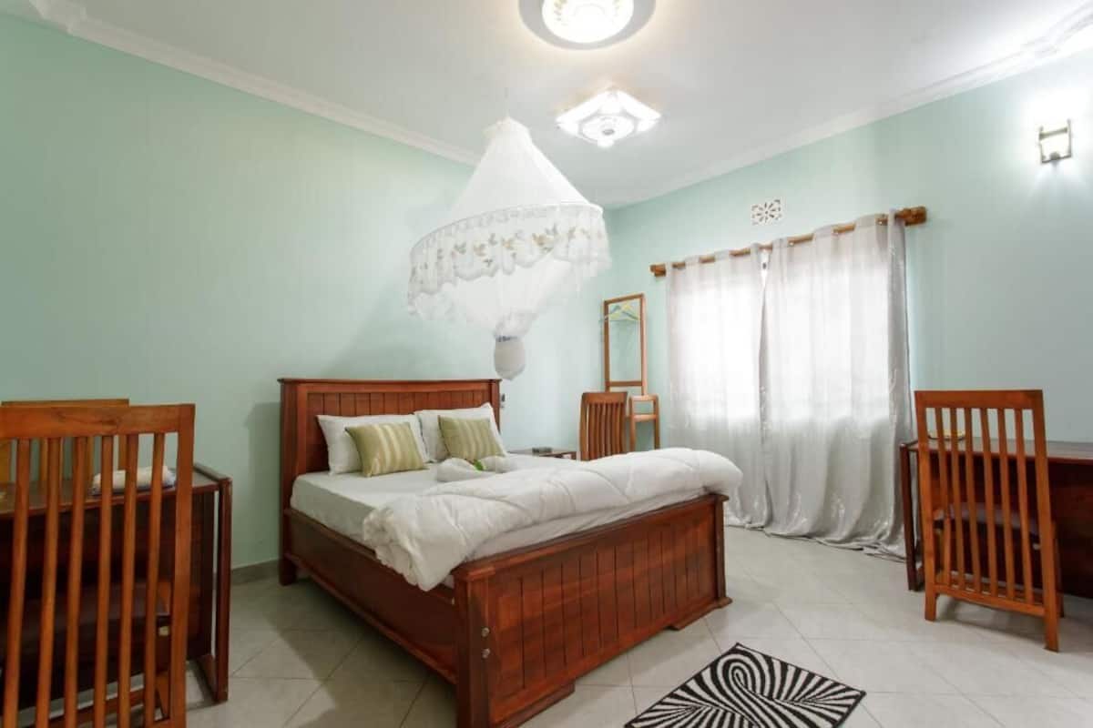 A bedroom with a wooden bed, a bedside table, a chair, a desk, a mirror, and a hanging light fixture.